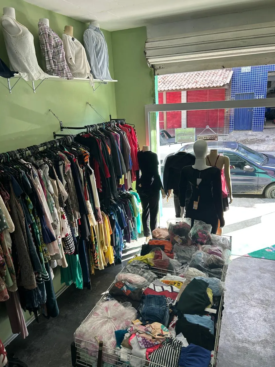 Interior do brechó com arara de roupas na parede, manequins na vitrine e cesto de peças em promoção